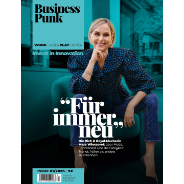 Frau sitzt mit einem Bein auf einem Sessel, trägt eine gepunktete Bluse mit Puffärmeln und Jeans, im Hintergrund ein Raum mit Steinwand und Holzbalken