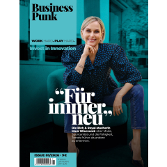 Frau sitzt mit einem Bein auf einem Sessel, trägt eine gepunktete Bluse mit Puffärmeln und Jeans, im Hintergrund ein Raum mit Steinwand und Holzbalken